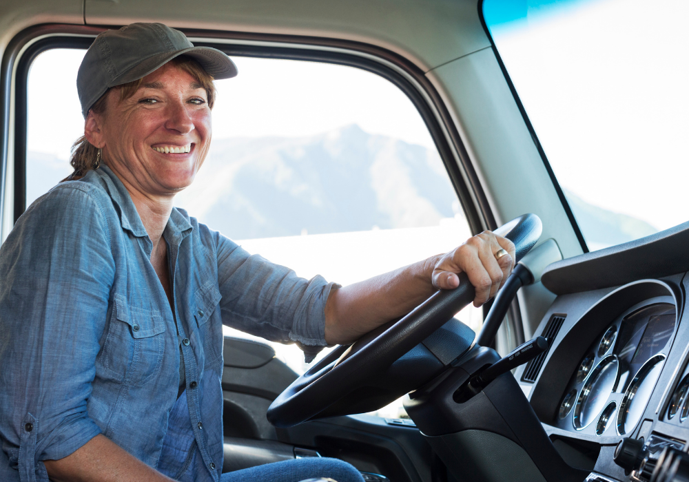 Female truck driver smiling while seated in the vehicle, showcasing a professional and friendly approach in the industry.